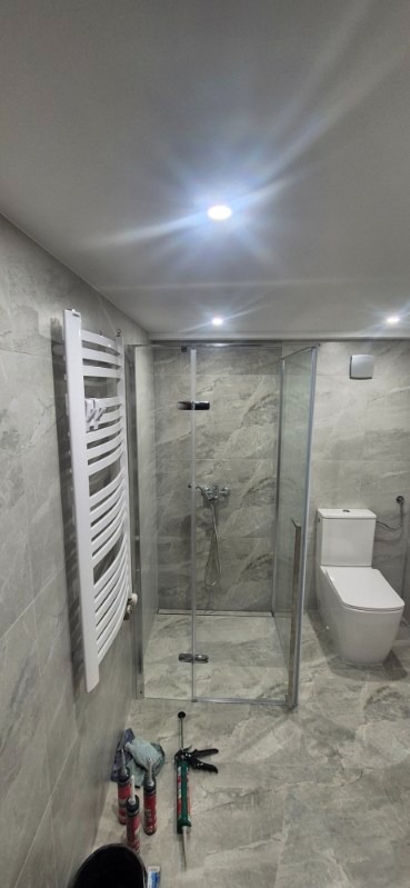 Wykończona łazienka z kabiną prysznicową typu walk-in, grzejnikiem drabinkowym i toaletą. Widoczne narzędzia i silikony sugerują świeży montaż.