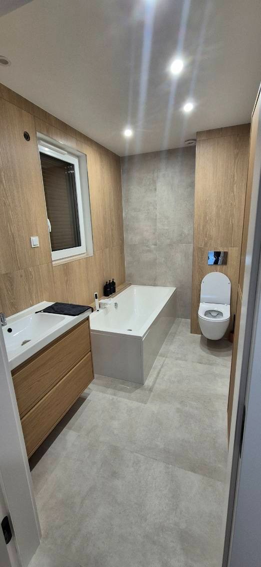 Nowoczesna łazienka z drewnianymi panelami, szarą płytką i białym wyposażeniem: wanna, umywalka z szafką i podwieszana toaleta. Oświetlenie punktowe.