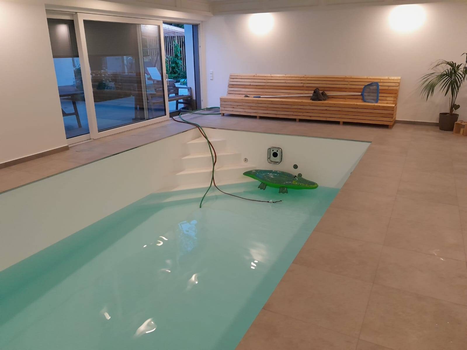 Wnętrze z basenem: jasne płytki podłogowe, białe ściany, drewniana ławka. W basenie pływa dmuchany krokodyl. Widoczne schody wejściowe i akcesoria do czyszczenia.
