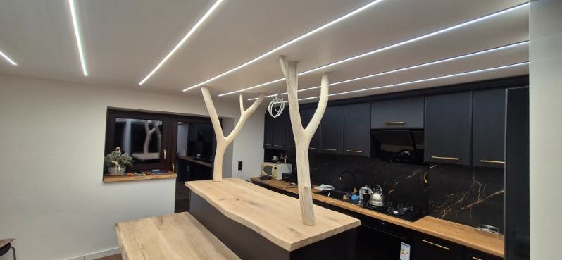 Nowoczesna kuchnia z czarnymi szafkami, blatem z efektem marmuru i wyspą z drewnianym blatem wspartą na białych, stylizowanych pniach drzew. Oświetlenie sufitowe LED.