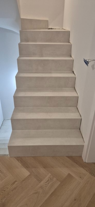 Schody wykończone jasnoszarym gresem, widok z dołu na drewnianą podłogę ułożoną w jodełkę. Ściana biała, widoczne niedociągnięcia przy wykończeniu.