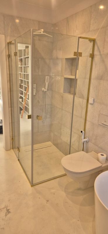 Nowoczesna kabina prysznicowa z elementami w kolorze złotym, marmurowe płytki, toaleta i fragment umywalki w jasnej łazience. Widok na regał z książkami przez szybę.