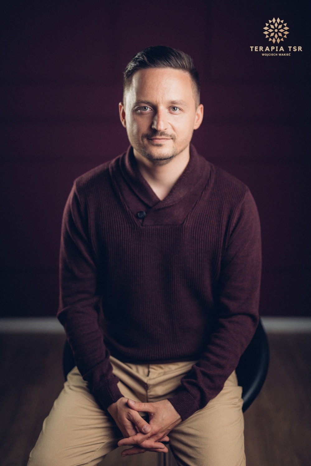 Portret mężczyzny w bordowym swetrze siedzącego na krześle z rękami złożonymi na kolanach, na tle śliwkowej ściany, z logo 'Terapia TSR Wojciech Wakieć' w prawym górnym rogu.