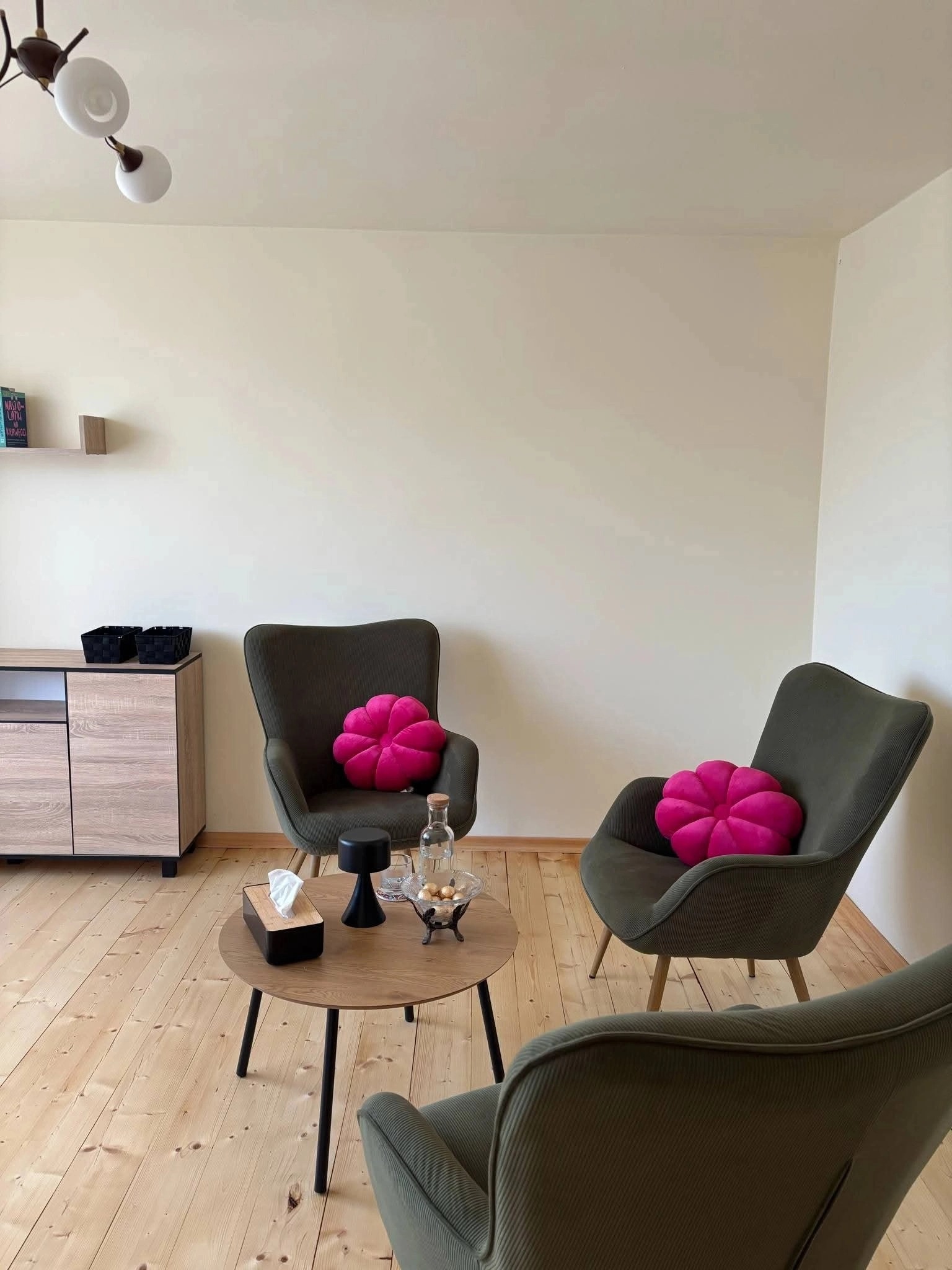 Jasne wnętrze gabinetu z trzema fotelami w odcieniach zieleni, okrągłym stolikiem z wodą i chusteczkami. Na fotelach różowe poduszki w kształcie kwiatów.