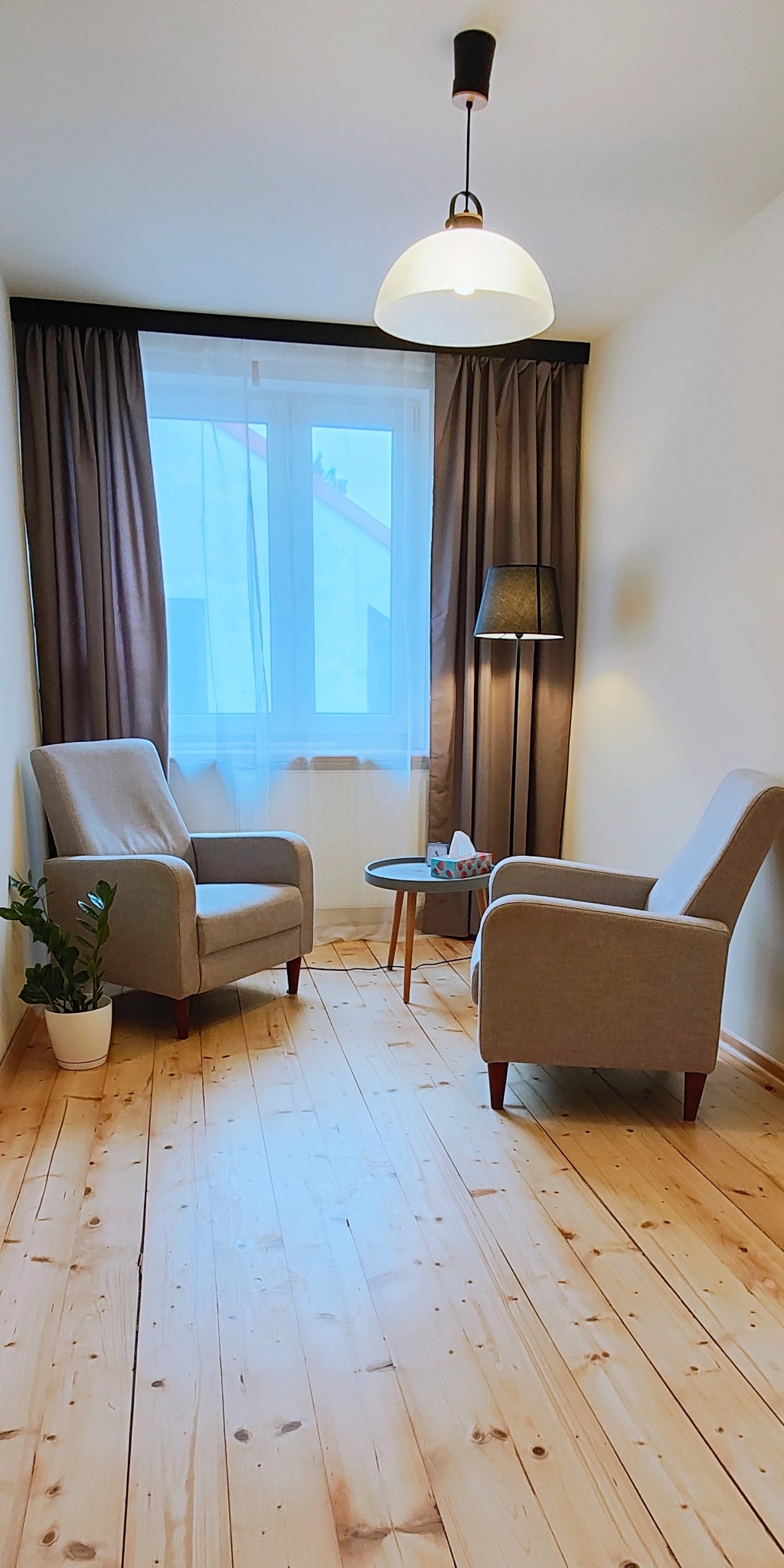 Jasny gabinet z dwoma fotelami, stolikiem i lampą podłogową. Drewniana podłoga, okno z zasłonami. Neutralna kolorystyka, spokojna atmosfera.