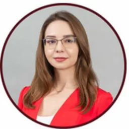 Portret kobiety w okularach, ubrana w czerwoną marynarkę, na neutralnym szarym tle.