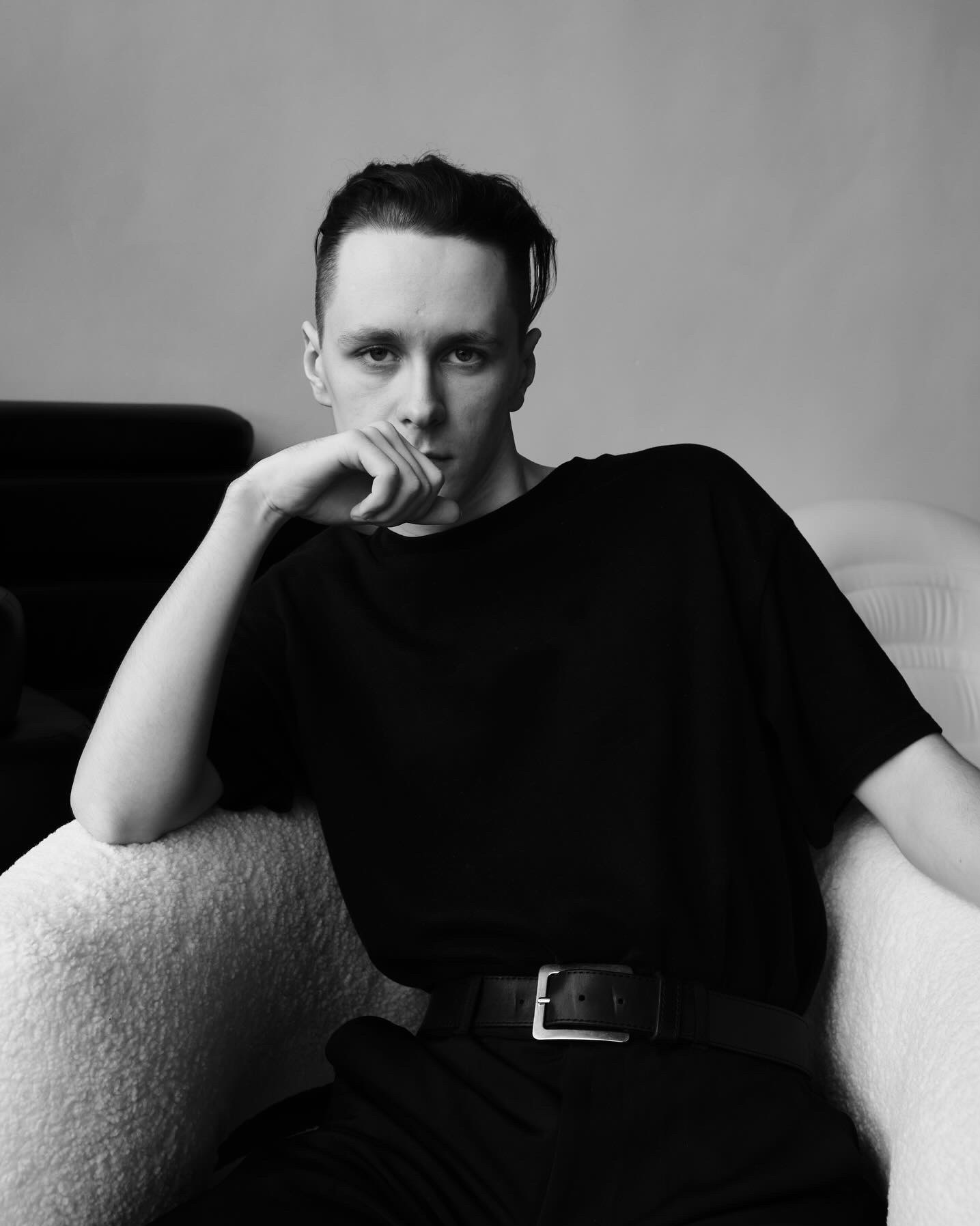 Portret młodego mężczyzny w czarnej koszulce i spodniach, siedzącego w fotelu o jasnym obiciu, z ręką podpartą o brodę, w czarno-białej tonacji.