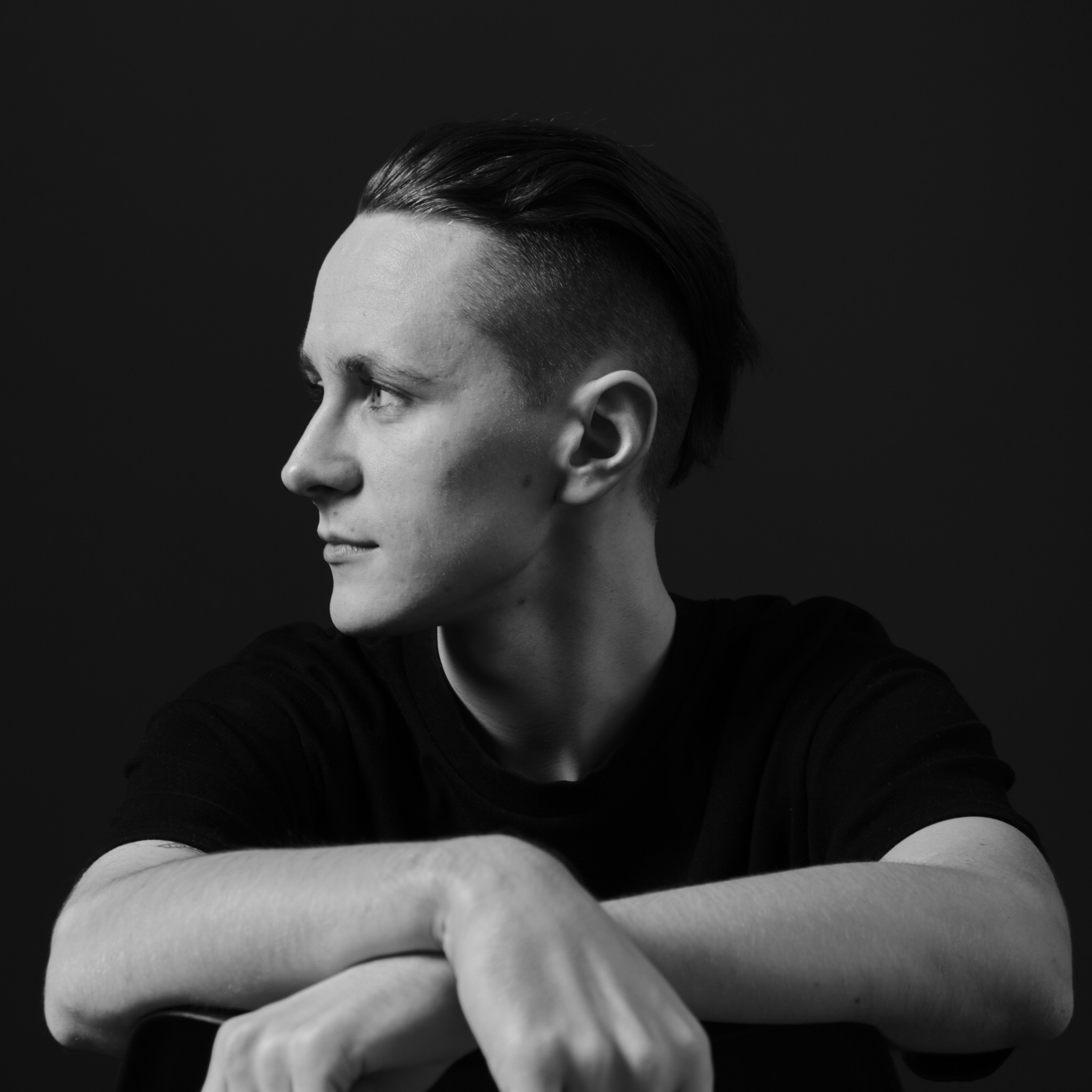 Czarno-białe, profilowe ujęcie młodego mężczyzny z modnym cięciem włosów, sfotografowanego w studiu w Warszawie, z rękami skrzyżowanymi na oparciu krzesła, na ciemnym tle.