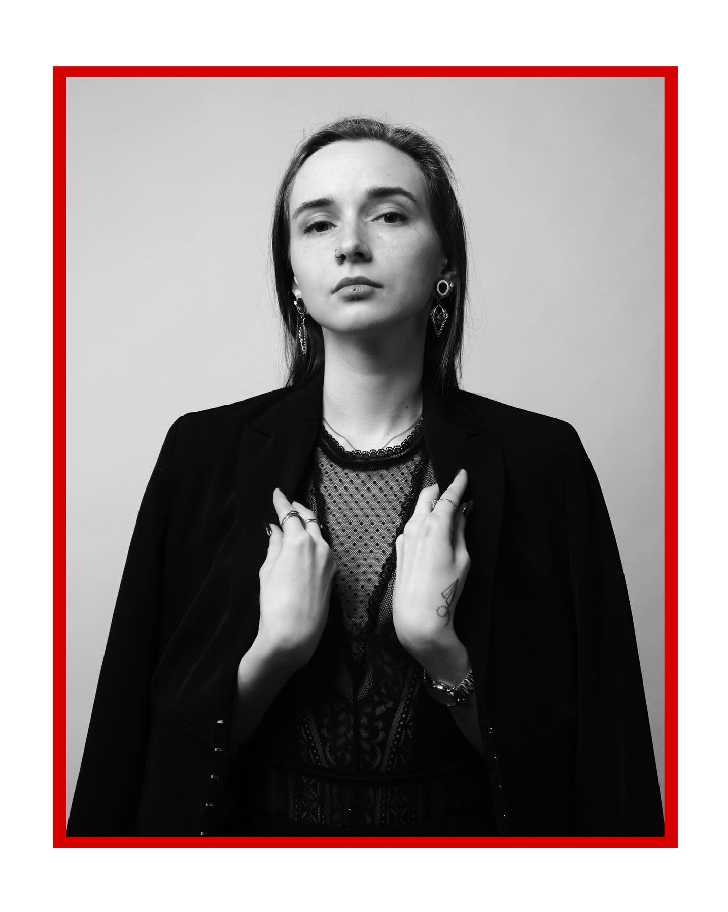 Portret kobiety w czarnej marynarce i koronkowej bluzce, z piercingiem w wardze i kolczykami, ręce na klapach marynarki, czarno-białe zdjęcie z czerwonym obramowaniem.
