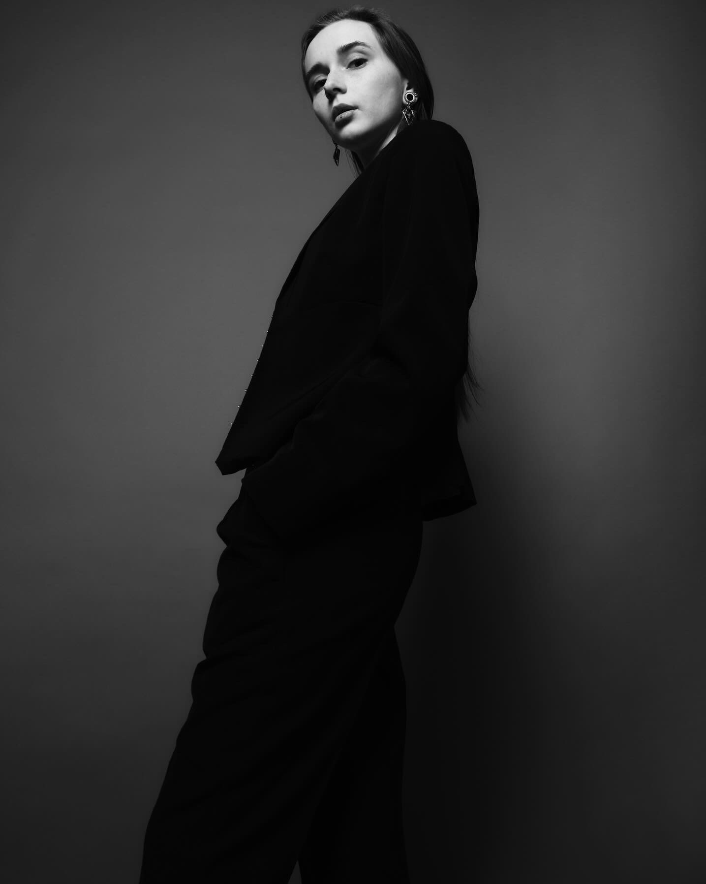 Czarno-białe zdjęcie kobiety w czarnym garniturze, z długimi włosami i kolczykami, pozującej na szarym tle.