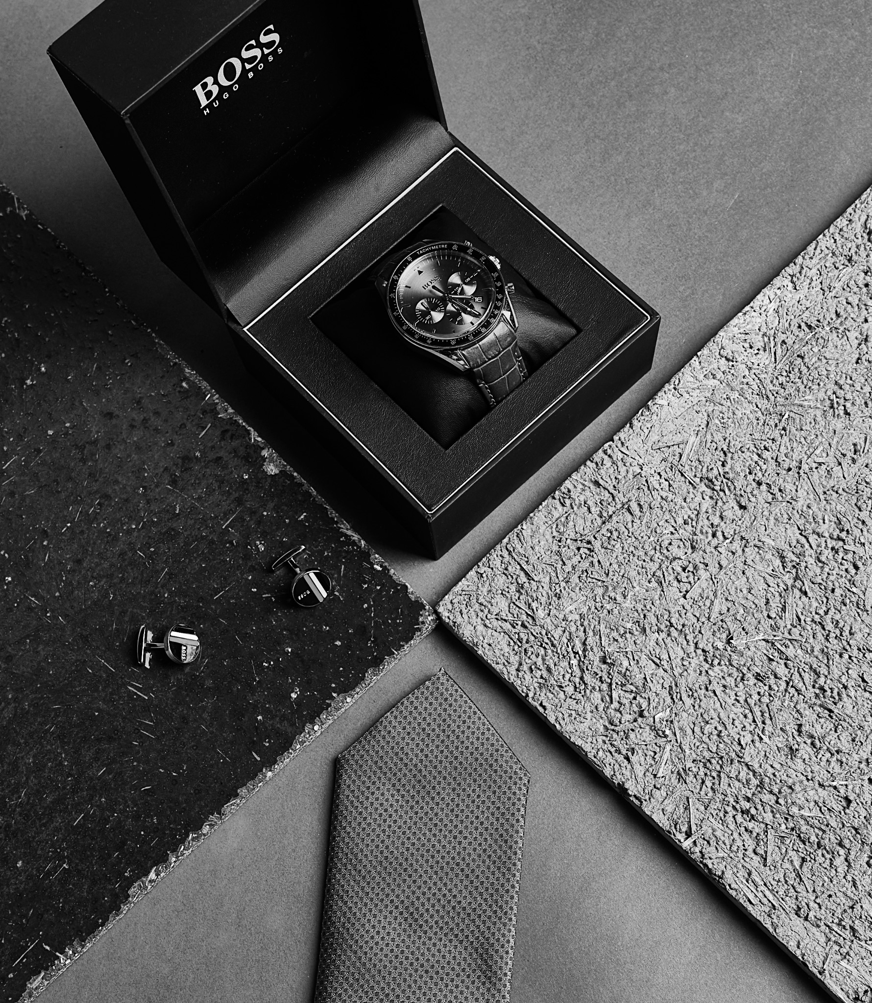 Czarno-białe ujęcie zegarka w pudełku marki Boss, spinek do mankietów i krawata na tle tekstur