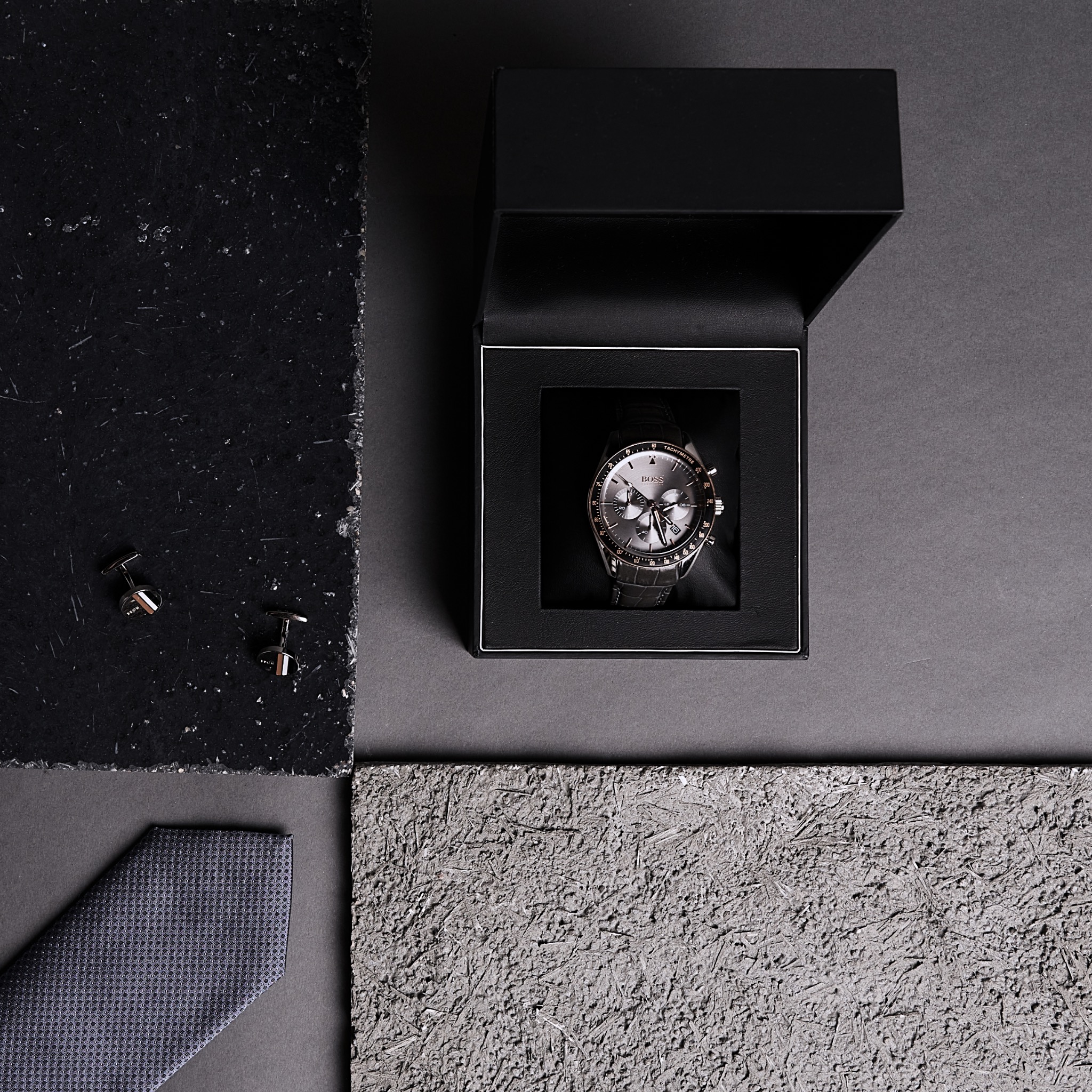 Zegarek męski marki BOSS w czarnym pudełku, spinki do mankietów i krawat na tle z teksturą kamienia i betonu.