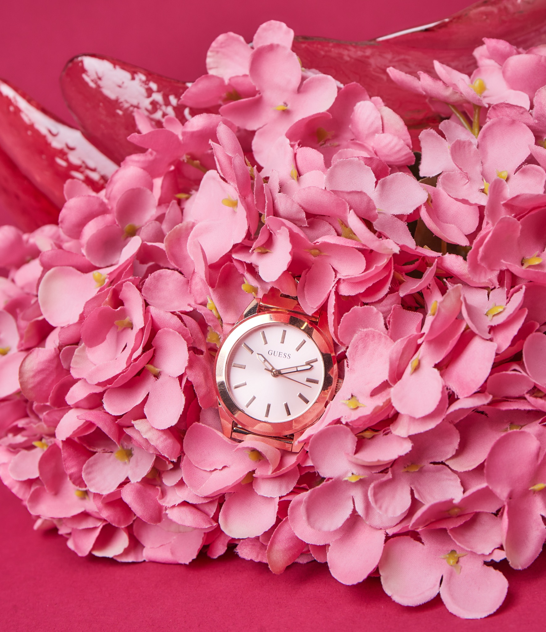 Zegarek damski marki Guess z różowozłotą bransoletą i białą tarczą, otoczony różowymi kwiatami na jednolitym różowym tle.