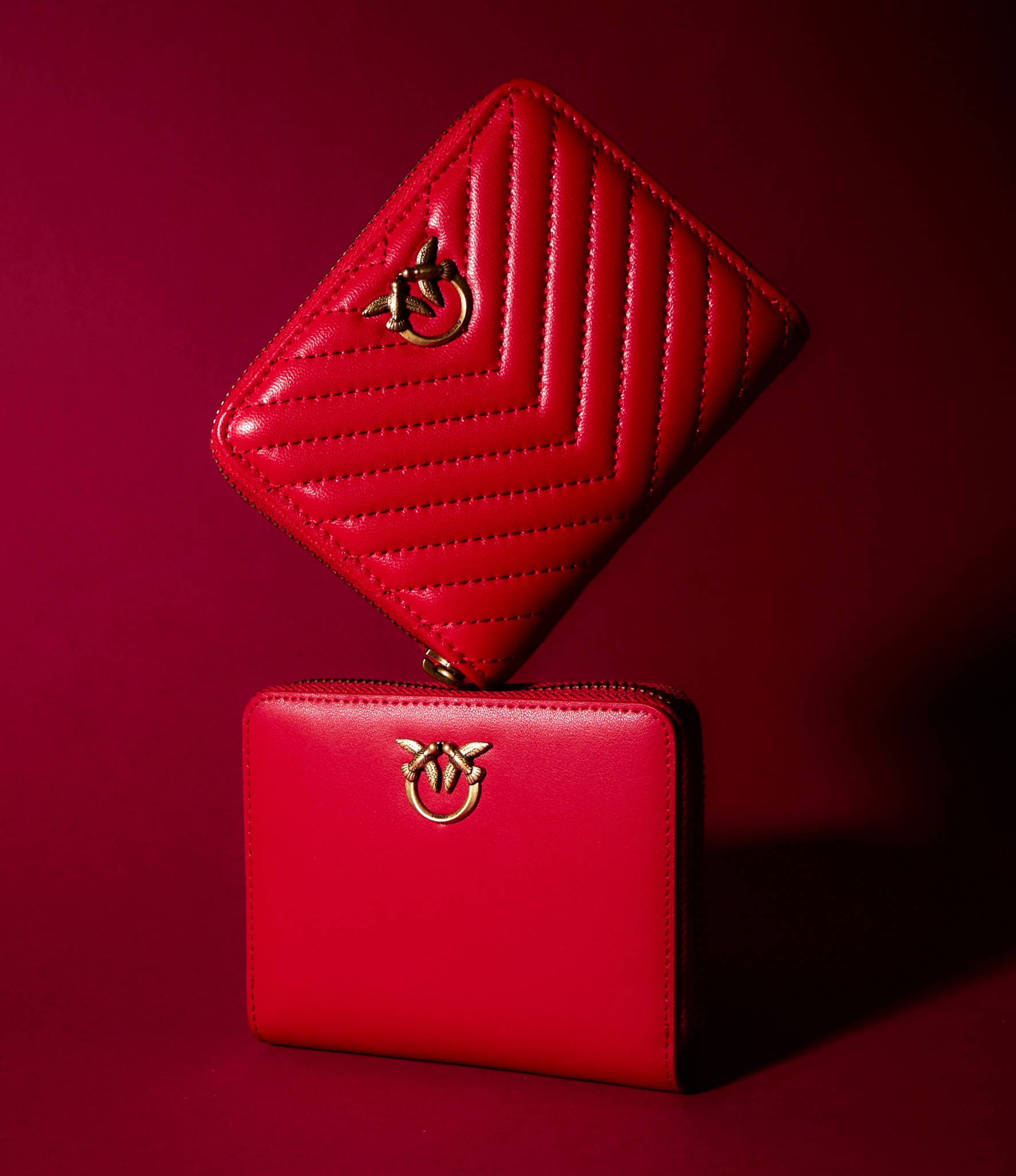 Dwa czerwone portfele Pinko na czerwonym tle, jeden z pikowaniem w jodełkę, drugi gładki, oba z logo z ptakami