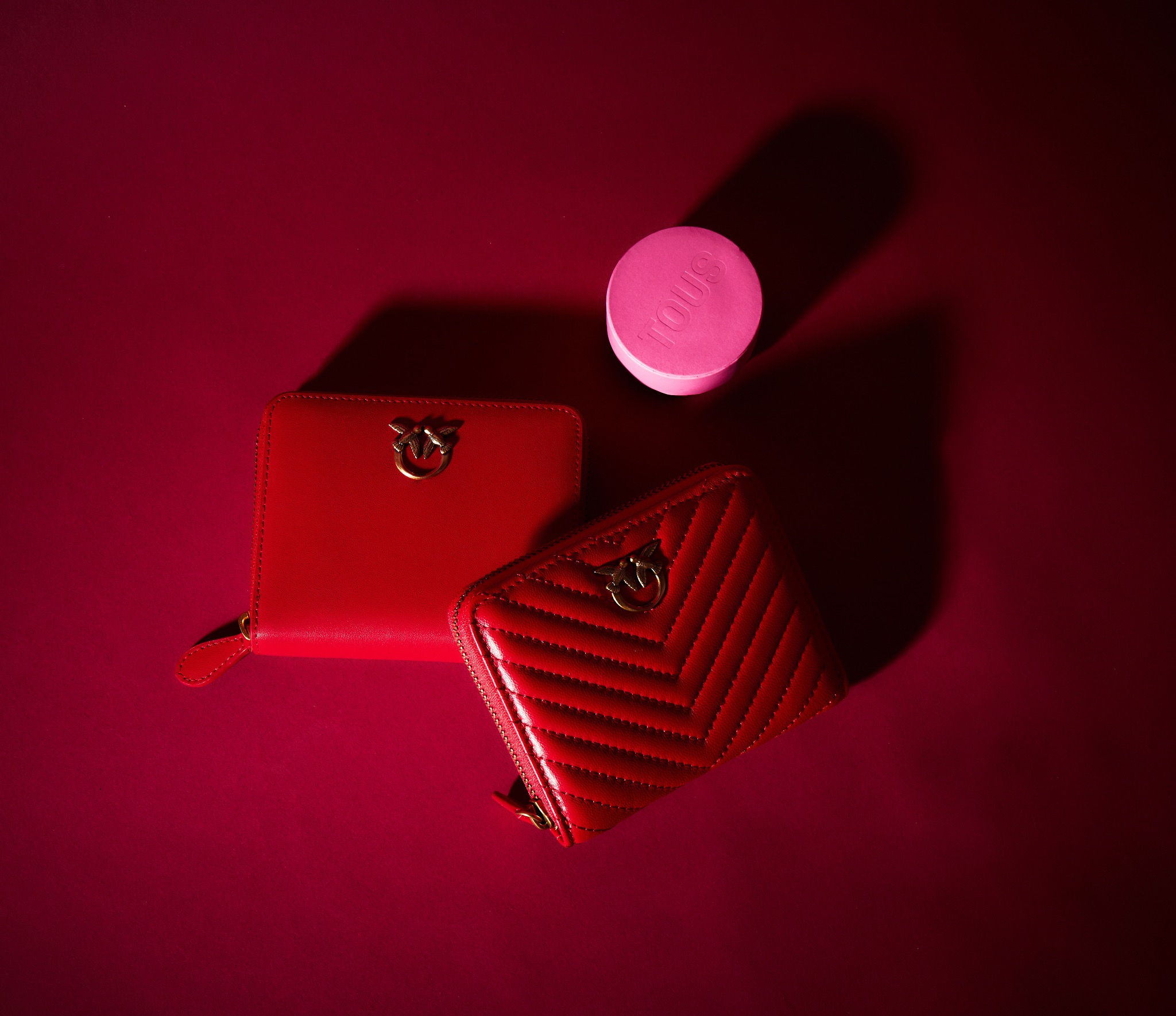 Dwa czerwone portfele damskie z logo Pinko leżące obok różowego, okrągłego pudełka Tous na tle bordowego tła.