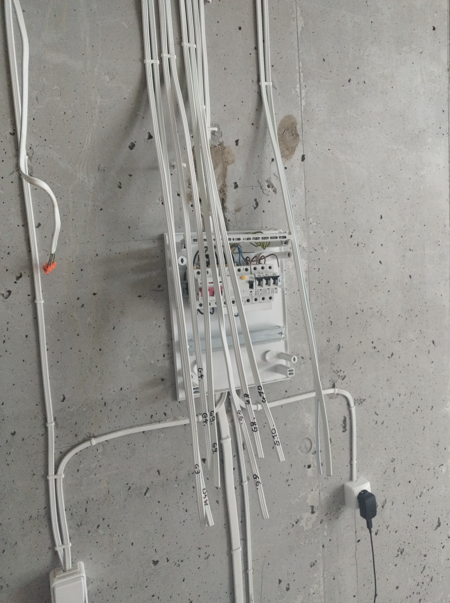 Szara betonowa ściana z zamontowaną białą skrzynką rozdzielczą, z której zwisają opisane kable; widoczne także gniazdko elektryczne z wtyczką, przewody ułożone wzdłuż ściany i przytwierdzone...
