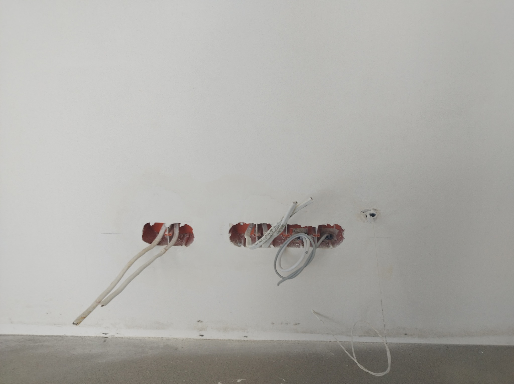 Przygotowane otwory w ścianie z wystającymi przewodami elektrycznymi, widoczne puszki instalacyjne w kolorze pomarańczowym, surowy stan wykończenia wnętrza.
