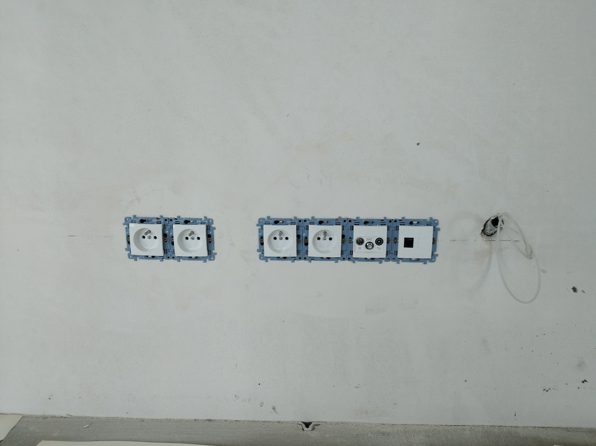 Surowa ściana z zamontowanymi puszkami podwójnego gniazdka elektrycznego, gniazdka antenowego RTV/SAT oraz gniazdka sieciowego, z widocznymi przewodami w otworze obok.