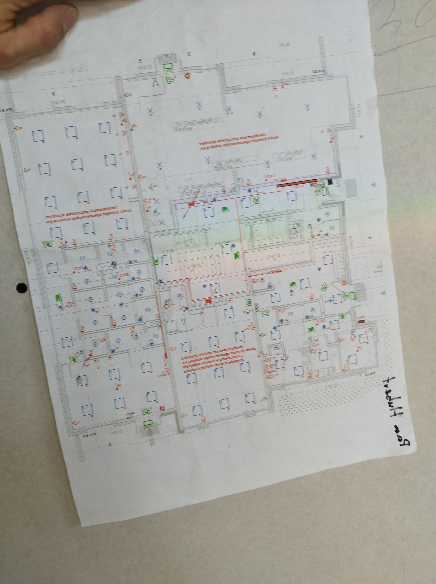 Plan instalacji elektrycznej lokalu, z naniesionymi punktami oświetleniowymi, gniazdkami i innymi elementami, widoczny fragment ręki w lewym górnym rogu.