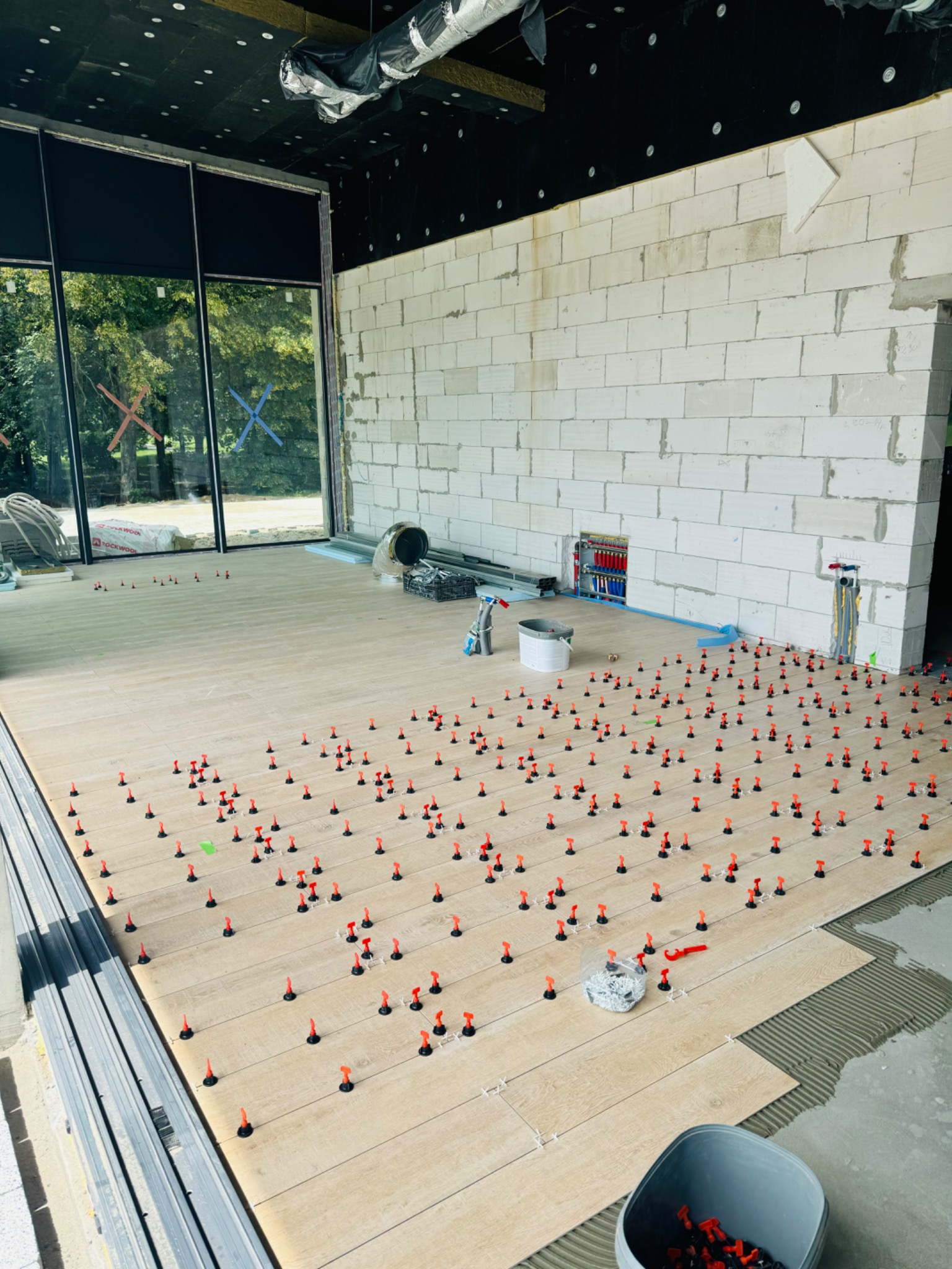 Układanie jasnych płytek podłogowych imitujących drewno w surowym wnętrzu z widocznymi elementami instalacji i dużą ilością czerwonych klinów poziomujących, z widokiem na zewnątrz przez duże okno.