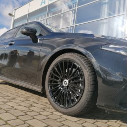 Świat felg i opon - Błyszczący, czarny Mercedes-Benz A-Klasa z czarnymi felgami aluminiowymi, zaparkowany na brukowanej nawierzchni przed nowoczesnym budynkiem z szklaną fasadą, widok z dołu.