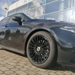 Błyszczący, czarny Mercedes-Benz A-Klasa z czarnymi felgami aluminiowymi, zaparkowany na brukowanej nawierzchni przed nowoczesnym budynkiem z szklaną fasadą, widok z dołu.