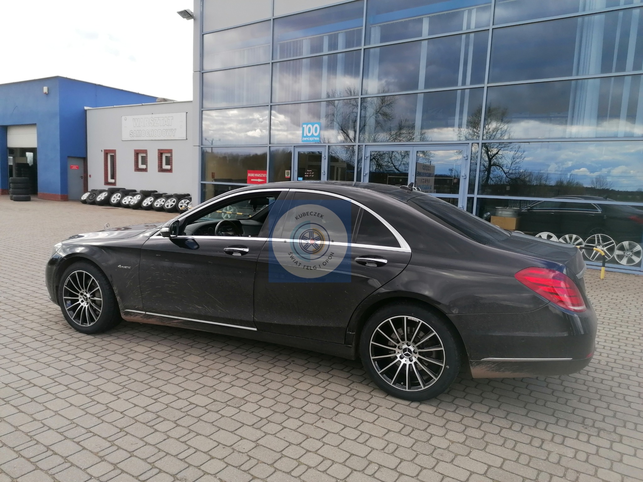 Czarny Mercedes klasy S, lekko zabrudzony, stoi przed budynkiem warsztatu samochodowego z oknami odbijającymi niebo.
