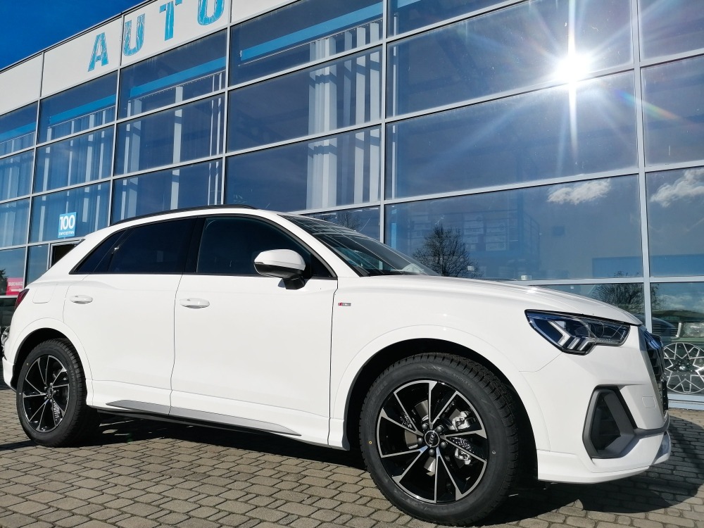 Białe Audi Q3 zaparkowane przed salonem samochodowym z dużymi szklanymi oknami, odbijającymi niebo i słońce.