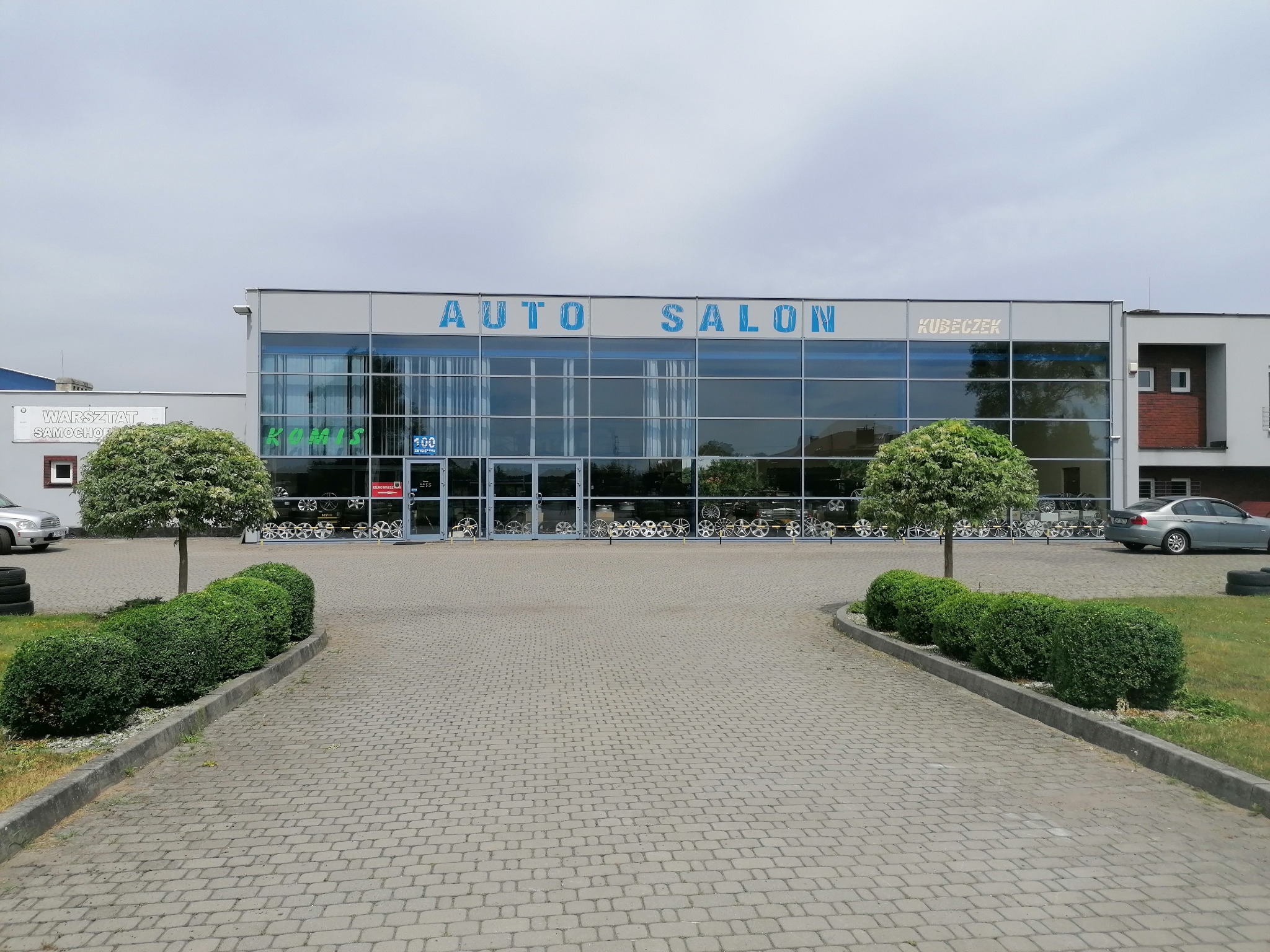 Fasada budynku Auto Salon Kubeczek z napisem AUTO SALON w kolorze niebieskim, widoczne felgi w oknach wystawowych, chodnik wyłożony kostką brukową i ozdobne krzewy.
