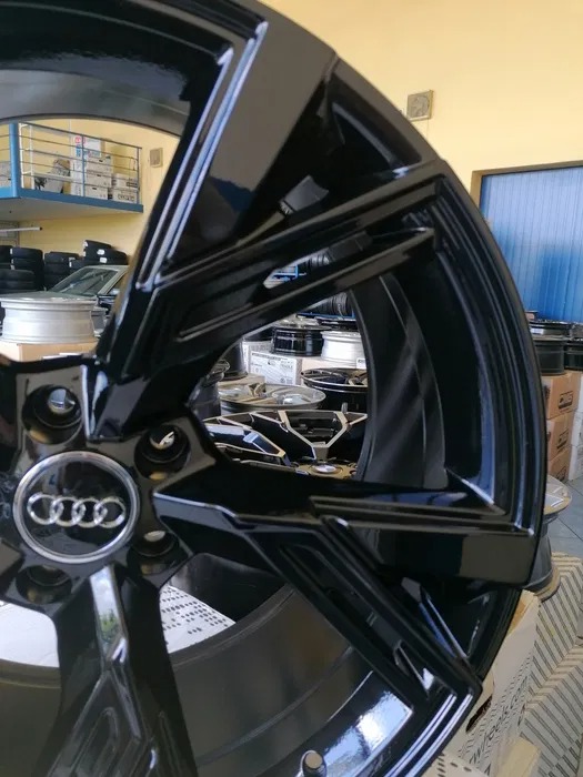 Błyszcząca, czarna felga aluminiowa Audi z widocznym logo, sfotografowana w warsztacie samochodowym na tle opon i pudeł.