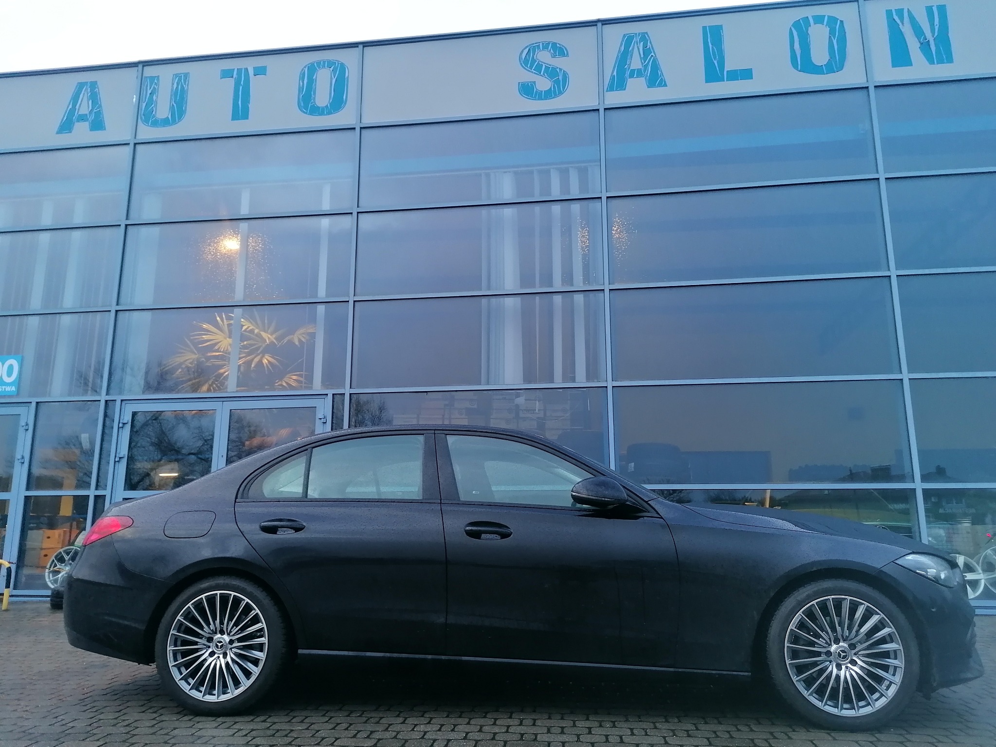 Czarny sedan marki Mercedes-Benz zaparkowany przed przeszklonym budynkiem z napisem 'AUTO SALON' w tle, widoczne felgi aluminiowe o skomplikowanym wzorze.