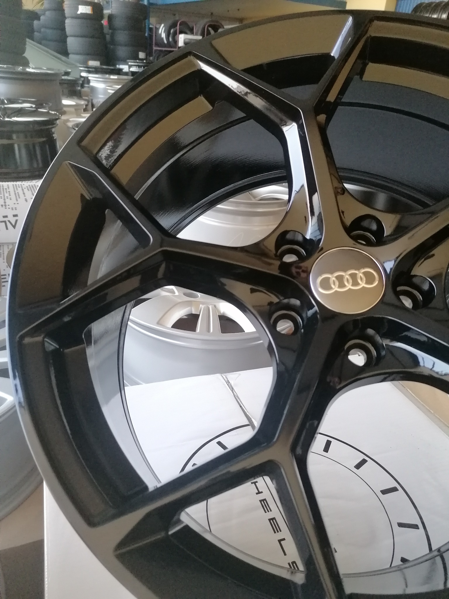 Błyszcząca, czarna felga aluminiowa Audi z widocznym logo na tle innych felg i opon w warsztacie.