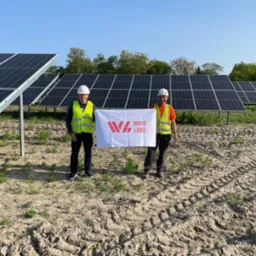 Dwaj mężczyźni w kaskach i kamizelkach odblaskowych trzymają biały baner z czerwonym logo firmy na tle rzędów paneli fotowoltaicznych na polu z widocznymi śladami opon.
