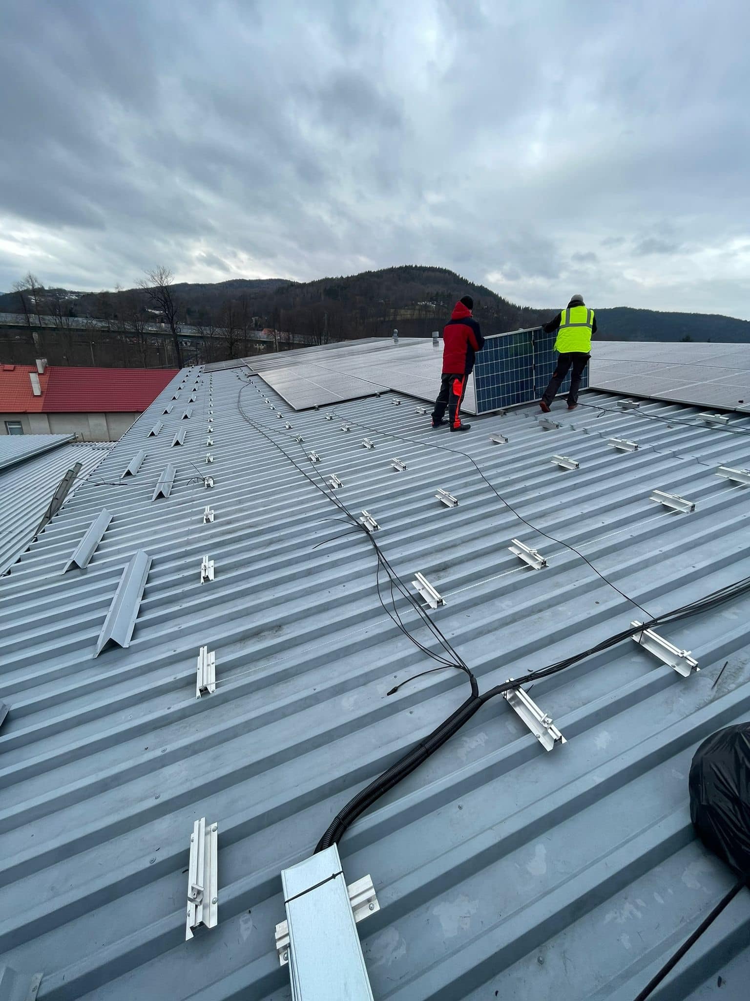 Montaż paneli fotowoltaicznych na metalowym dachu budynku przemysłowego, widoczne elementy mocujące i okablowanie, dwóch monterów w odzieży roboczej przenosi panel, pochmurne niebo w tle.