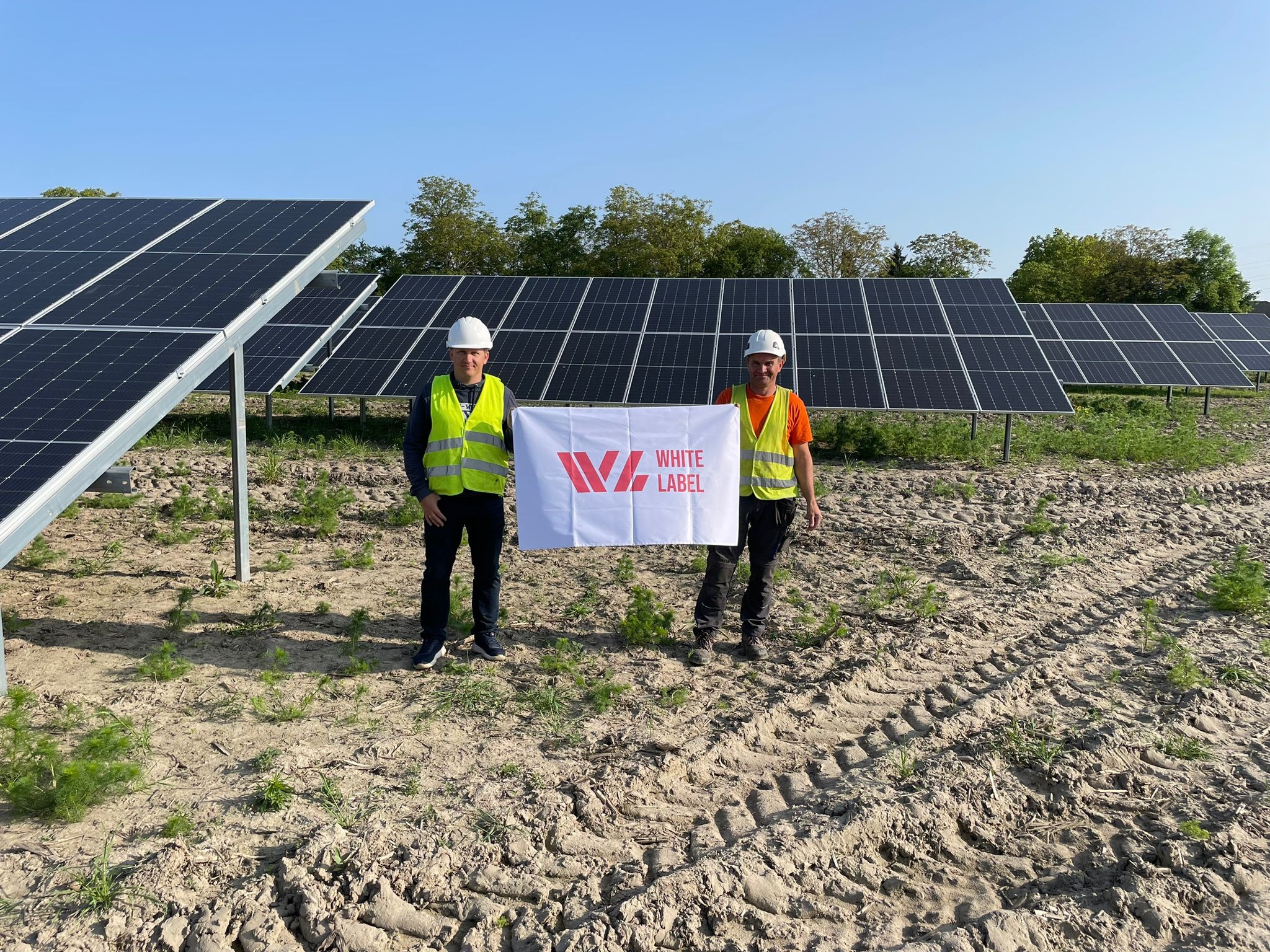 Dwaj mężczyźni w kaskach i kamizelkach odblaskowych trzymają biały baner z czerwonym logo firmy na tle rzędów paneli fotowoltaicznych na polu z widocznymi śladami opon.