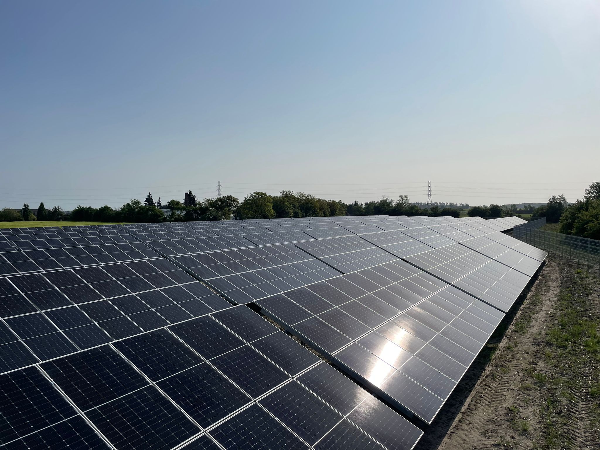 Rozległa farma paneli słonecznych pod błękitnym niebem, z widocznymi liniami wysokiego napięcia w oddali i śladami opon na ziemi.