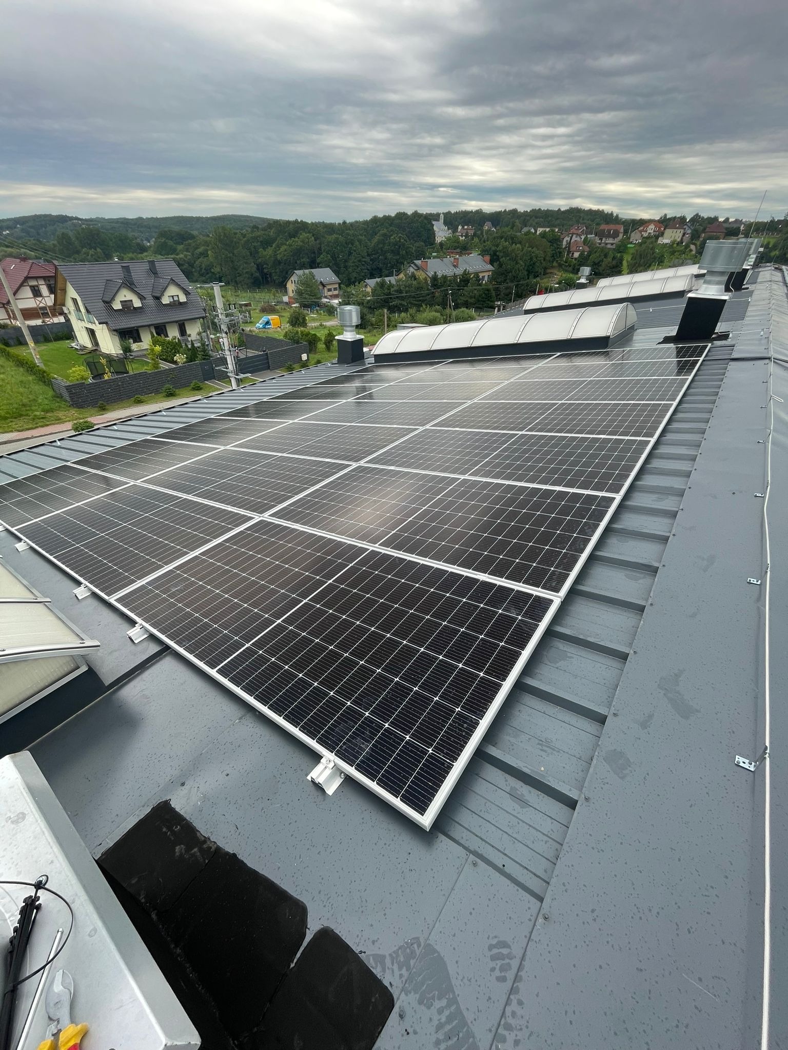 Panel słoneczny z widocznymi mocowaniami na szarym, metalowym dachu budynku przemysłowego; w tle zabudowa jednorodzinna i pochmurne niebo.
