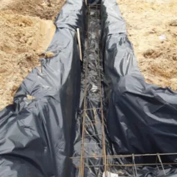 Wykonany wykop pod fundament, zabezpieczony czarną folią, zbrojenie stalowe w środku, widoczne ślady ziemi i piasku.