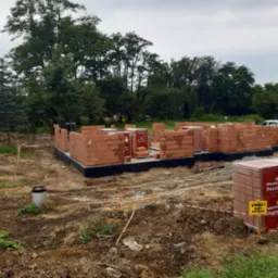 Wznoszenie murów parteru domu z czerwonej cegły Porotherm na fundamencie z izolacją przeciwwilgociową, widoczne palety z cegłą oraz tablica ostrzegawcza 'Uwaga! Roboty budowlane', w tle drzewa.