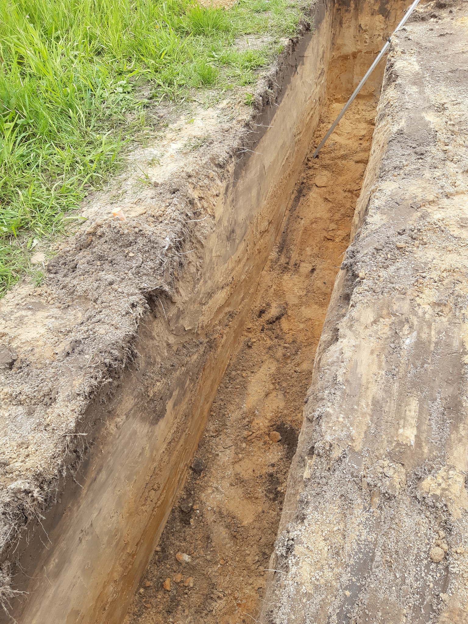 Wykop pod fundament z widoczną warstwą gruntu, śladami korzeni i metalową łatą wewnątrz wykopu.