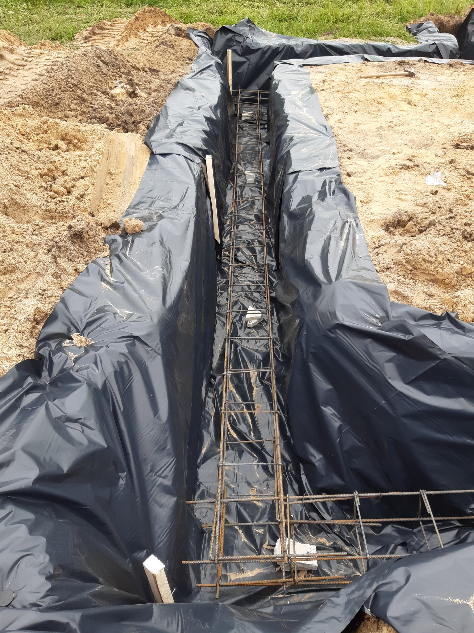 Wykonany wykop pod fundament, zabezpieczony czarną folią, zbrojenie stalowe w środku, widoczne ślady ziemi i piasku.