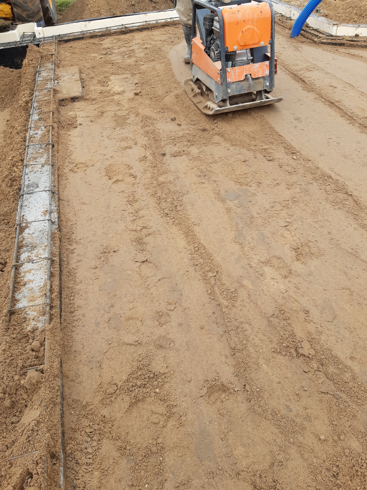 Pomarańczowa zagęszczarka wibrująca ubija piasek na budowie fundamentów, obok widoczny zbrojony betonowy wieniec.