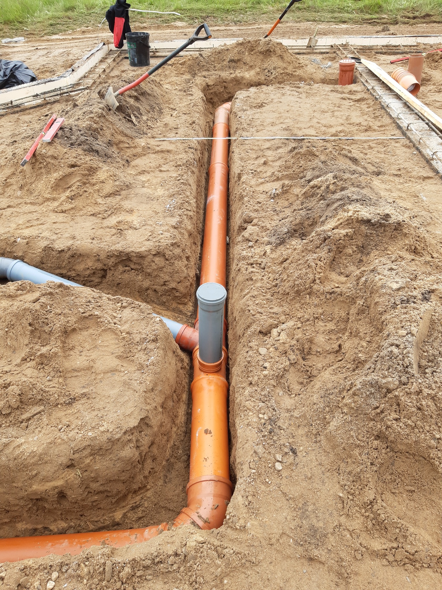 Pomarańczowe rury kanalizacyjne ułożone w wykopie, widoczne elementy szalunku i narzędzia budowlane w tle.