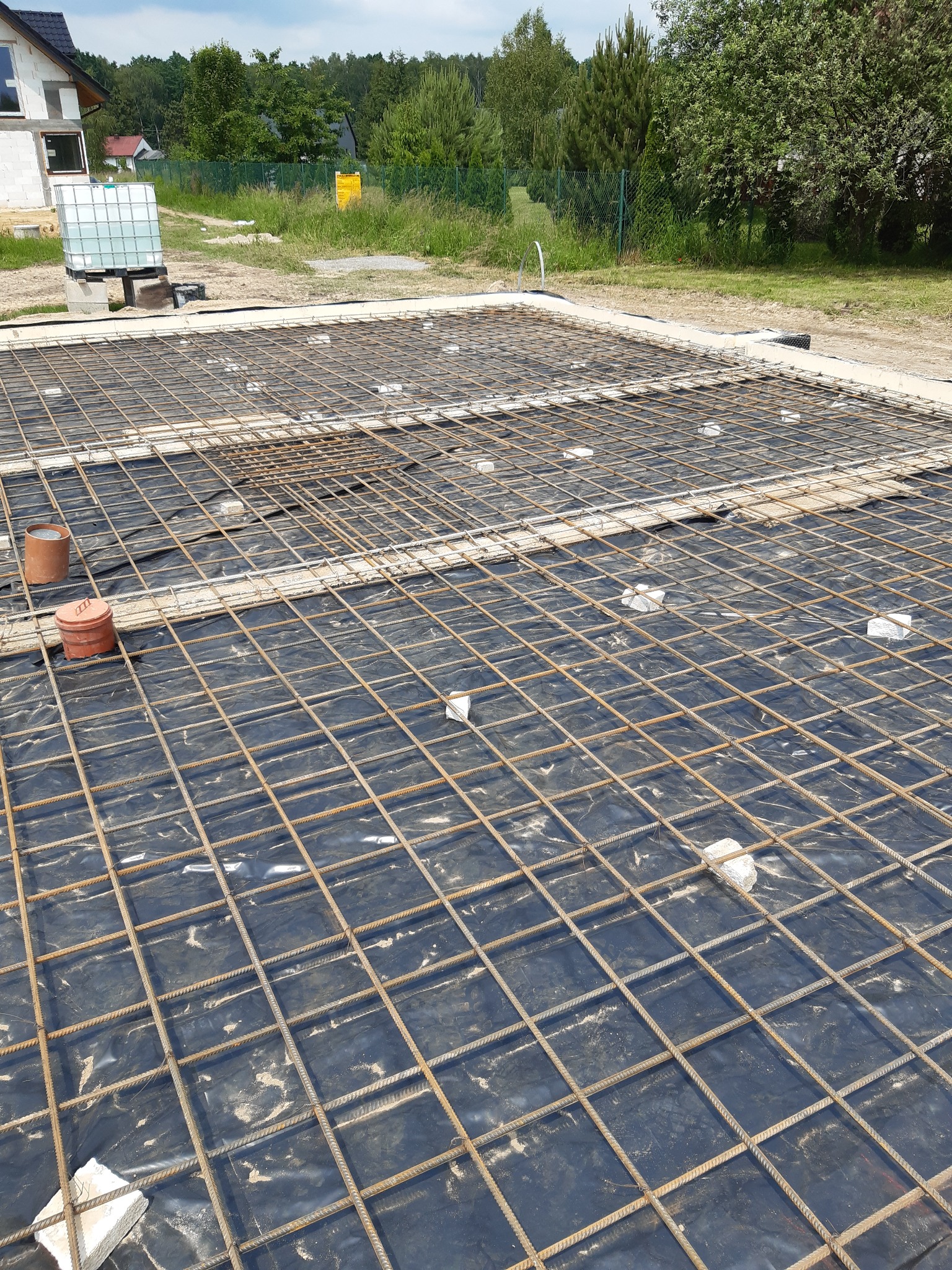 Zbrojenie fundamentu na czarnej folii z widocznymi elementami dystansowymi i rurami kanalizacyjnymi w tle plac budowy z budynkiem w stanie surowym otwartym.