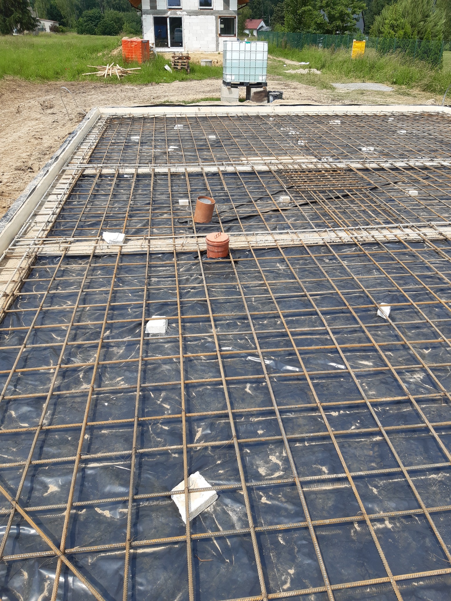 Zbrojenie płyty fundamentowej ułożone na czarnej folii, z widocznymi dystansami i szalunkiem, w tle budynek w stanie surowym otwartym.