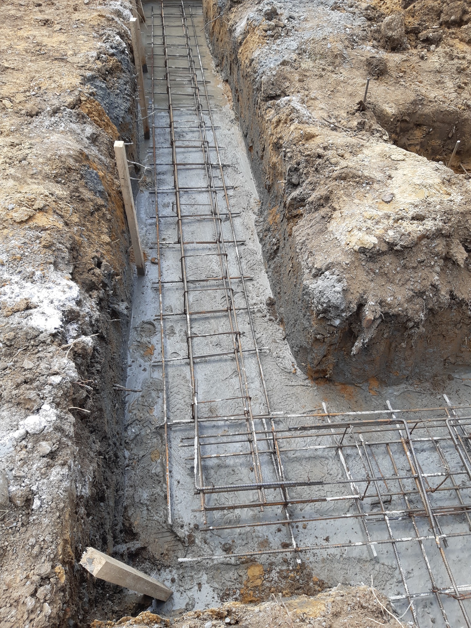 Wykop pod fundament z częściowo zalanym betonem i zbrojeniem z prętów stalowych, widoczne drewniane szalunki i ziemia.