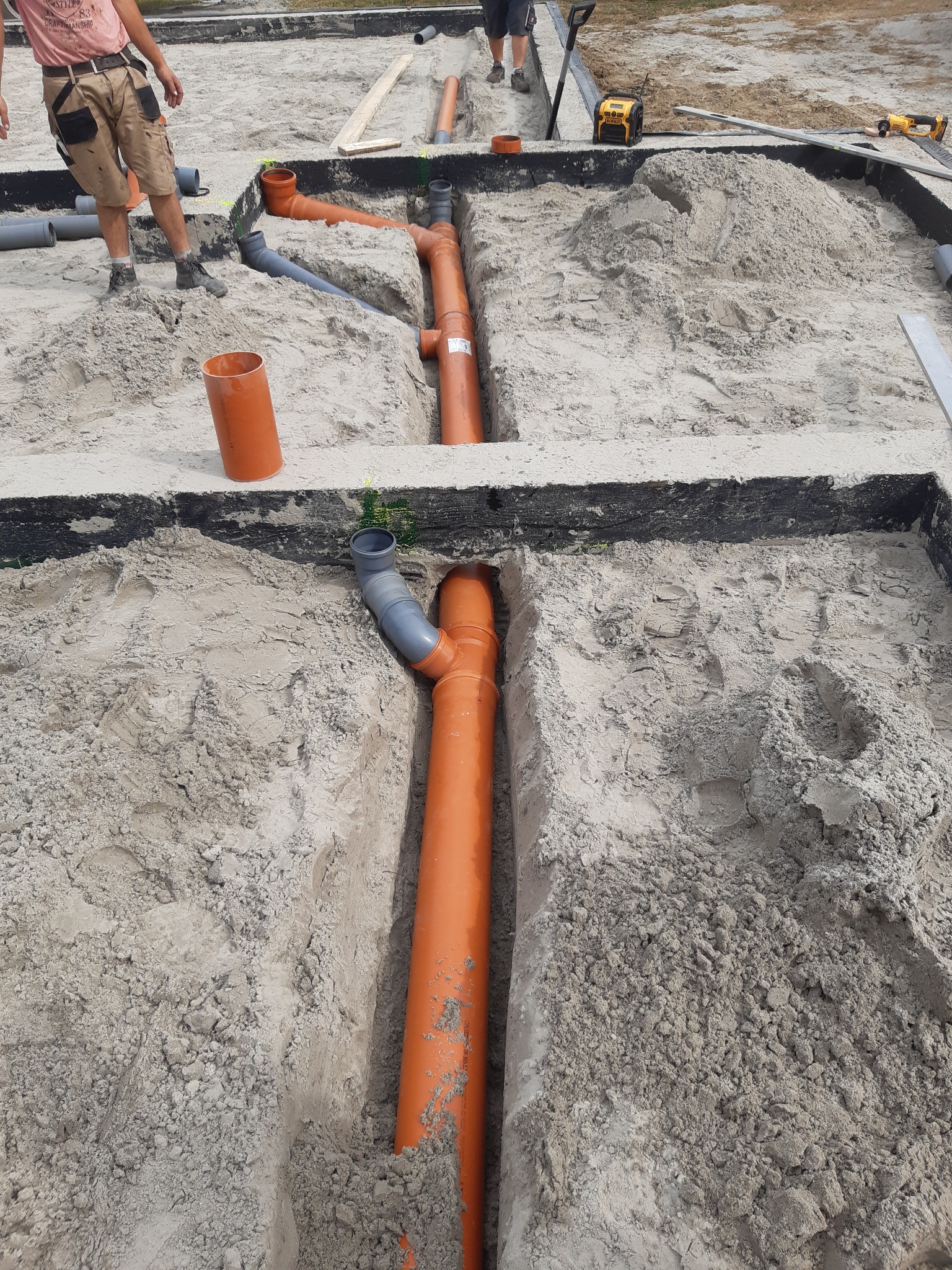 Pomarańczowe rury kanalizacyjne ułożone w wykopie fundamentowym, częściowo zasypane piaskiem, z widocznymi kształtkami i pracownikami w tle.