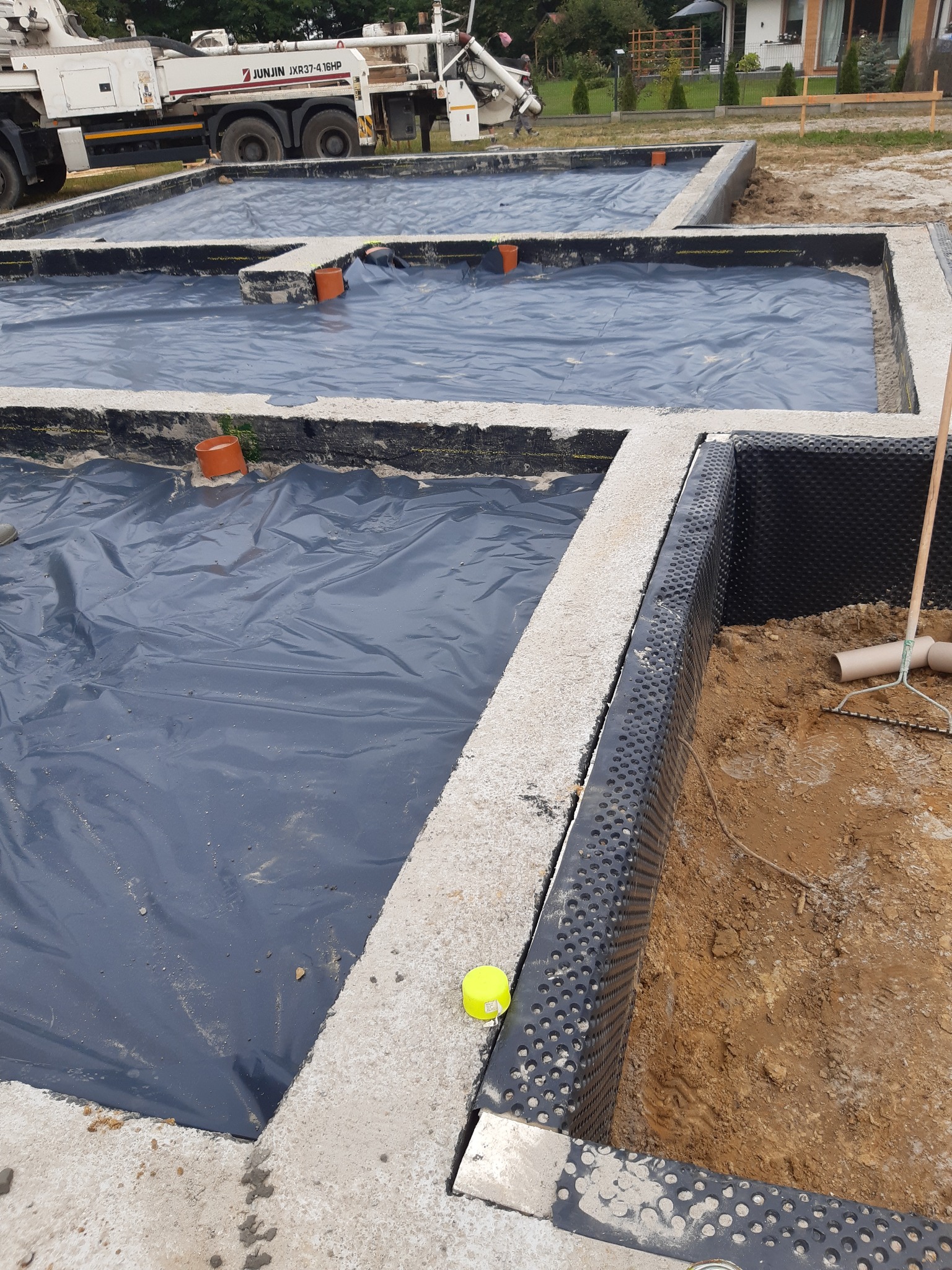 Wykop pod fundamenty zabezpieczony folią hydroizolacyjną i membraną kubełkową, z widocznym betonowym wieńcem i rurami kanalizacyjnymi, na tle betonomieszarki.