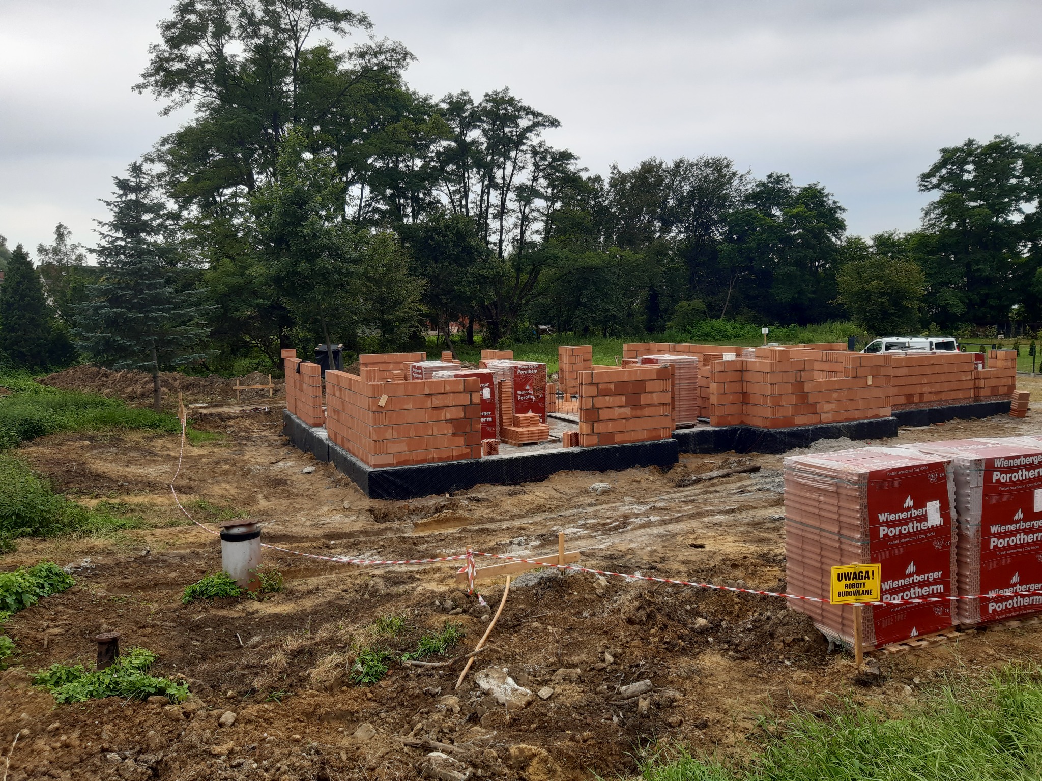 Wznoszenie murów parteru domu z czerwonej cegły Porotherm na fundamencie z izolacją przeciwwilgociową, widoczne palety z cegłą oraz tablica ostrzegawcza 'Uwaga! Roboty budowlane', w tle drzewa.