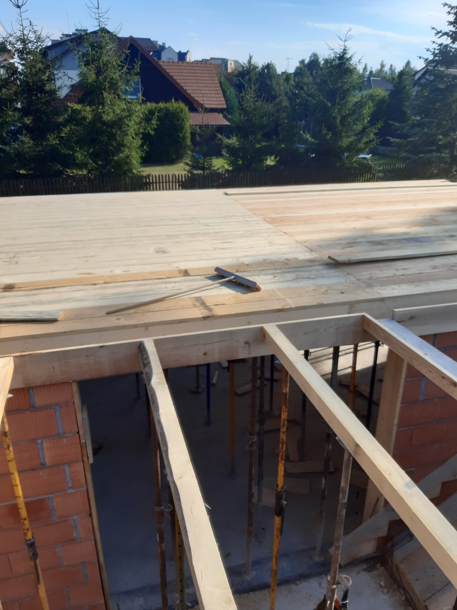 Wznoszenie ścian z czerwonej cegły i drewnianego szalunku pod strop; widoczne podpory stropu i częściowo ułożone drewniane deski stropowe, w tle zieleń drzew i zabudowa mieszkalna.
